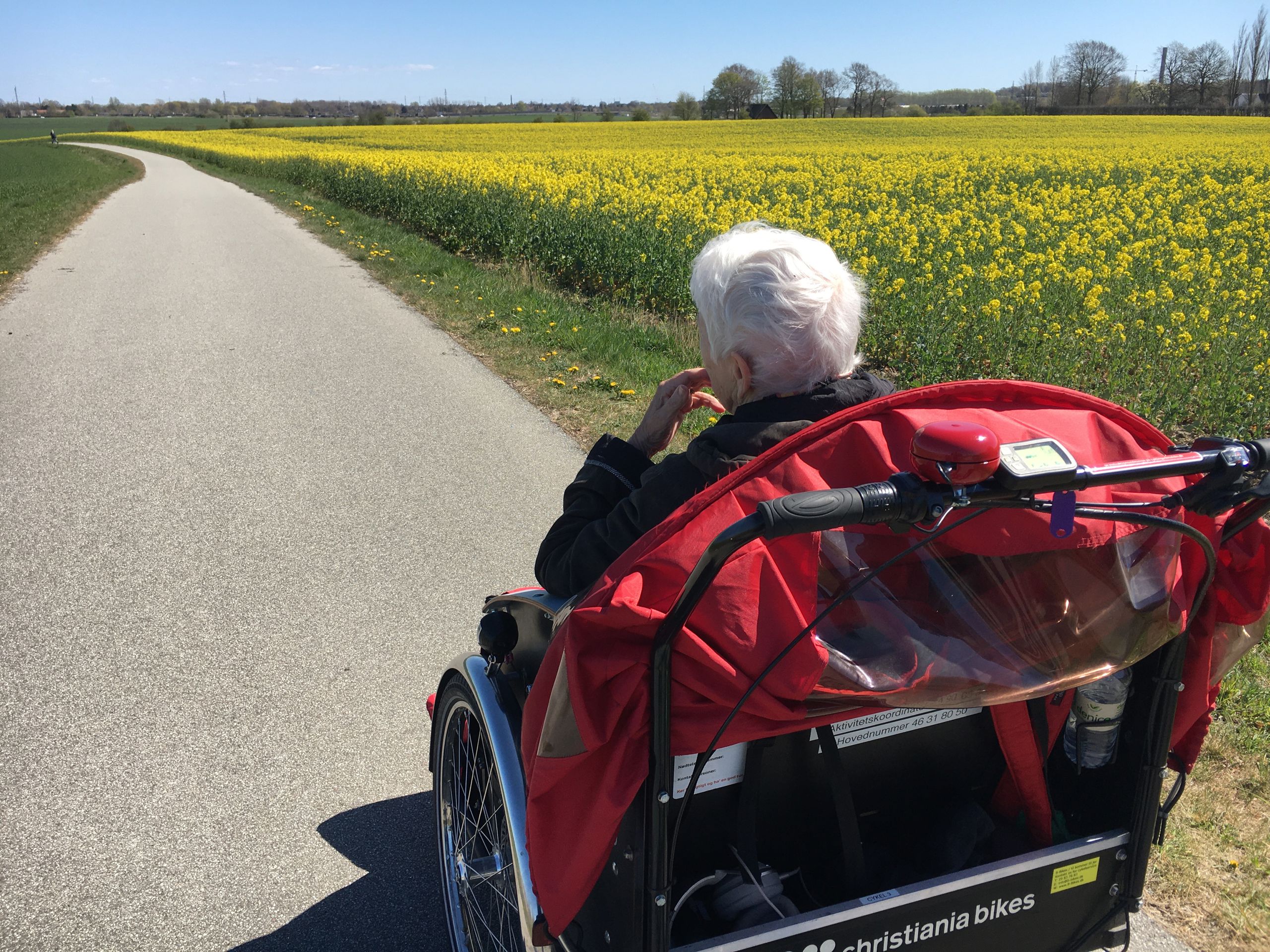Plejehjemsbeboer sidder i en rickshaw-cykel. De er ude i naturen, ved en gul rapsmark.